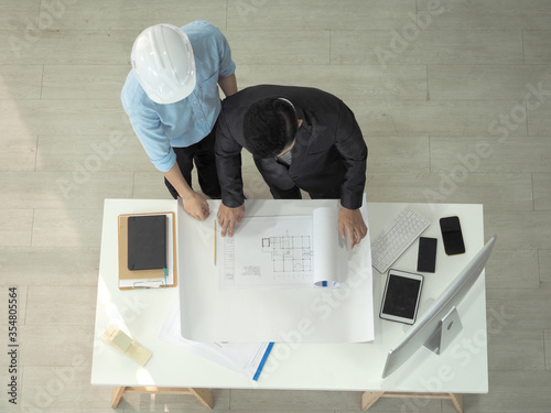 Top view of asian businessman and architect or engineer are discussing work together in office.Man pointing at blueprint.Foreman is wearing construction helmet.