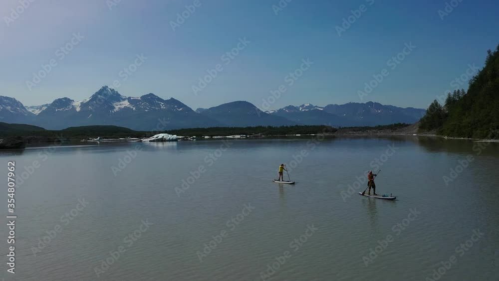 fly away from two paddle boarders on lake full of ice bergs and pan right past green forested mountain to show the Valdez glacier cascading down the mountains. fly away from two paddle boarders on lake full of ice bergs and pan right past green forested mountain to show the Valdez glacier cascading down the mountains.