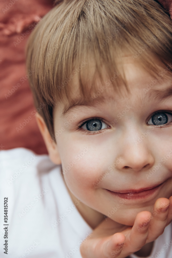 Soft focus. Portrait of adorable little boy with blond hairs and blue eyes, indoor. Boy is lying on a bed with terracotta bed linen on the background Soft focus. Portrait of adorable little boy with blond hairs and blue eyes, indoor. Boy is lying on a bed with terracotta bed linen on the background