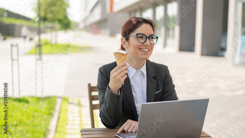 Smiling woman working remotely on a laptop and eating ice cream cone while sitting at a wooden table outdoors. Portrait of a girl in a business suit for a lunch break in a street cafe.