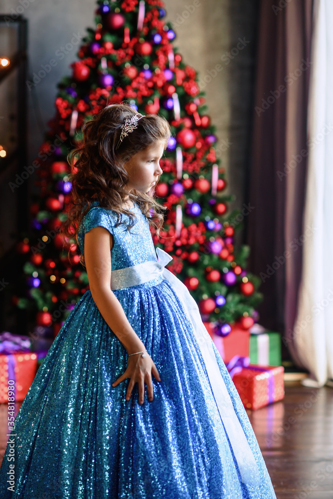 Fototapeta premium girl with dark hair standing on a box with gifts. Christmas tree in the background. smiles