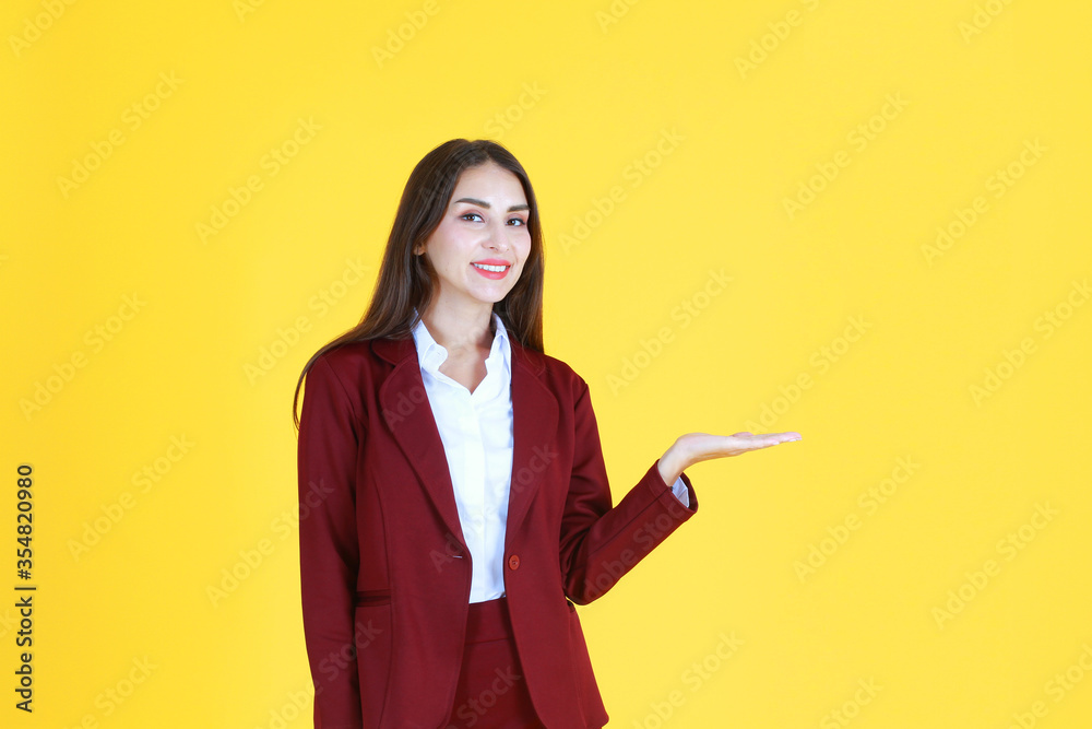 Smart Caucasian woman in red suit smiling and her hand open for present isolated on yellow background. Copy space Smart Caucasian woman in red suit smiling and her hand open for present isolated on yellow background. Copy space