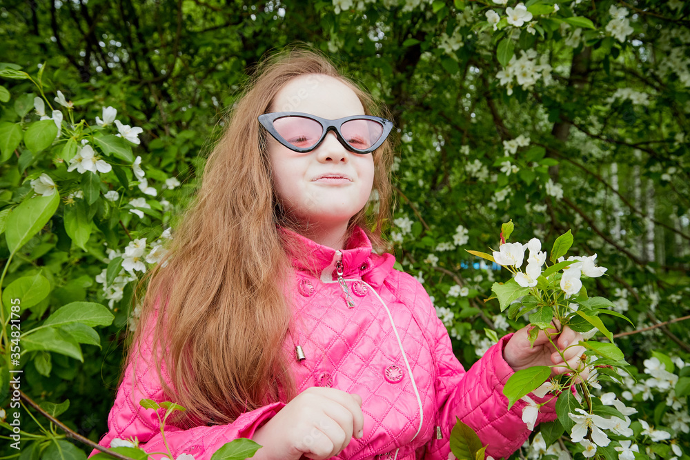 Pretty cute little girl with long hair and in pink glasses posing near a blooming apple tree with white flowers in a summer park
