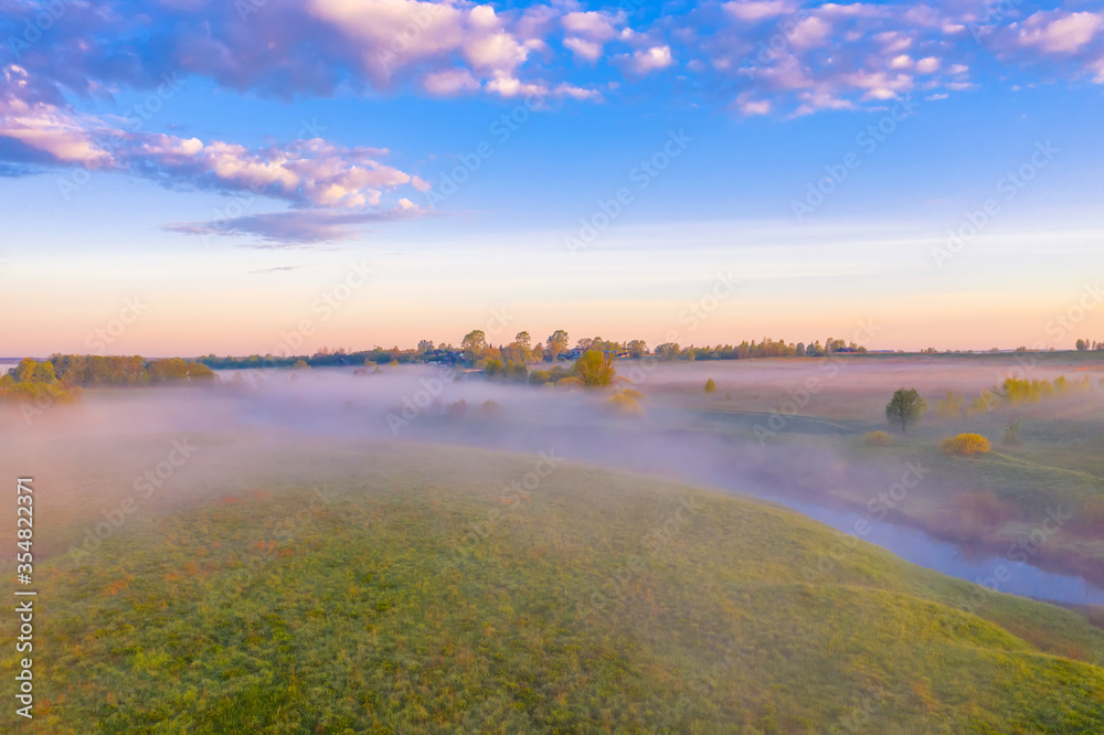 Morning fog in the meadows and at the mouth of the river during sunrise in the countryside, view from the height of the landscape on the horizon forest and village.