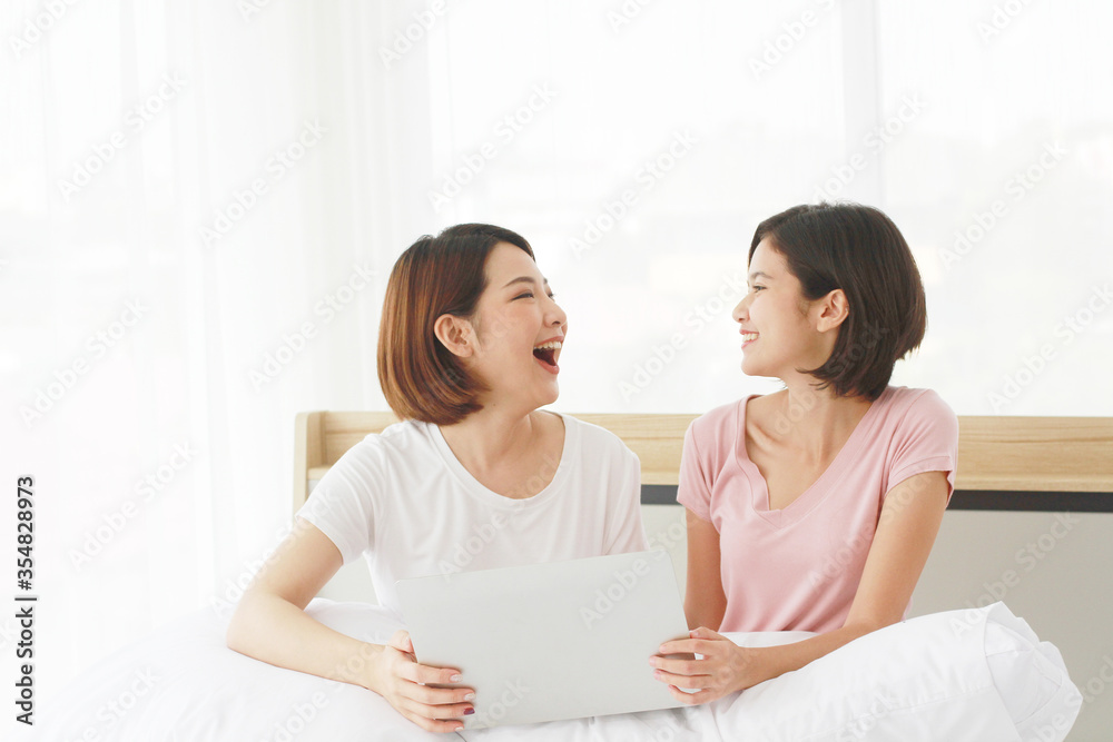 Asian young sisters lovely couple sitting on white bed and smiling together for online Social with laptop in bedroom. Homosexual women or Lesbian in love. LGBT Asian and Holiday concept. Asian young sisters lovely couple sitting on white bed and smiling together for online Social with laptop in bedroom. Homosexual women or Lesbian in love. LGBT Asian and Holiday concept.