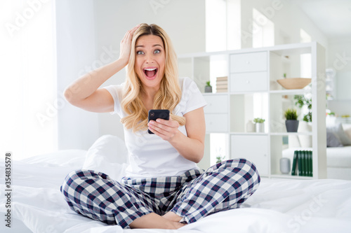 Photo of funny shocked lady hold telephone read news arm on head sitting bed freelancer quarantine stay home white linen legs crossed perfect sunny morning wear pajama room indoors