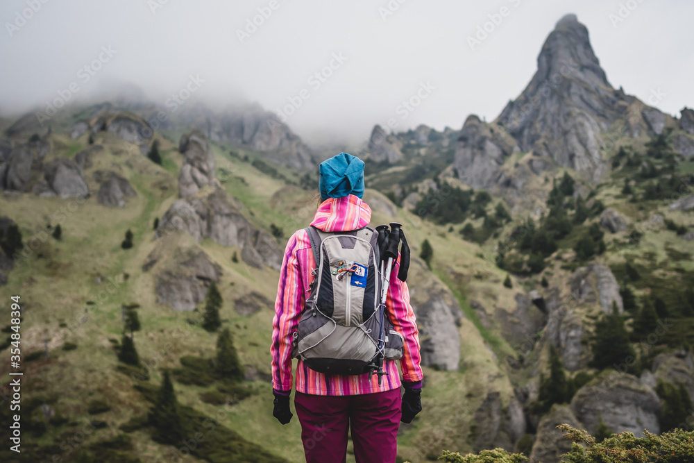 Successful female hiker with backpack on her back observing and admiring the mountain peak. Beautiful nature landscape with a young woman in hiking activity in a mountainous area of Romania. Successful female hiker with backpack on her back observing and admiring the mountain peak. Beautiful nature landscape with a young woman in hiking activity in a mountainous area of Romania.
