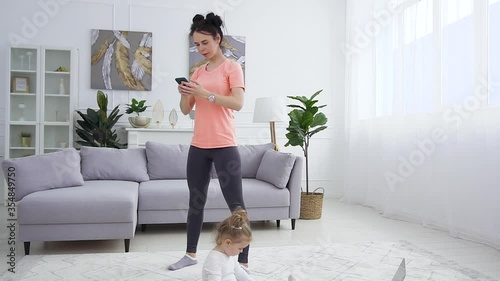 Joyful pretty sporty young woman in sportswear using mobile during her fitness squating exercises while her small nice daughter playing on the carpet photo