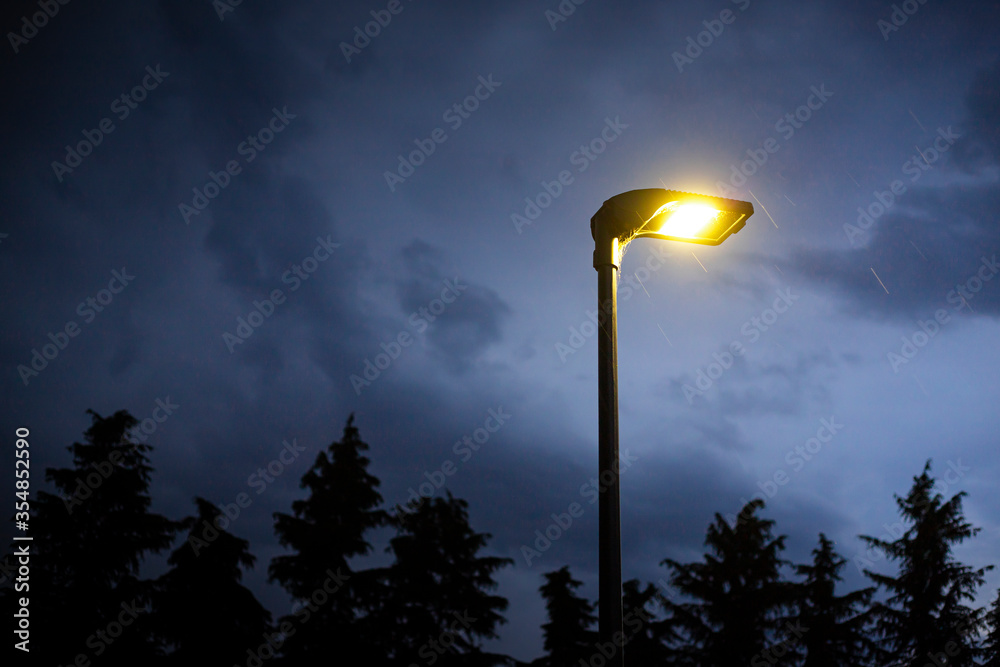 Lit street lamp at twilight with moody blue sky, trees silhouettes in the background and rain falling. Gloomy and spooky background with copy space on the left.