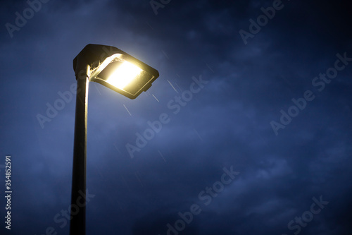 Lit street lamp at twilight with moody blue sky and rain falling. Gloomy and spooky background with copy space on the right.