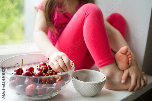 child girl sitting on windowsill at home and eating sweet cherries. summer fruits  healthy seasonal vitamin food