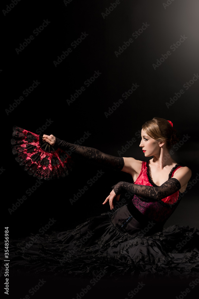 young woman holding fan while sitting and dancing flamenco on black