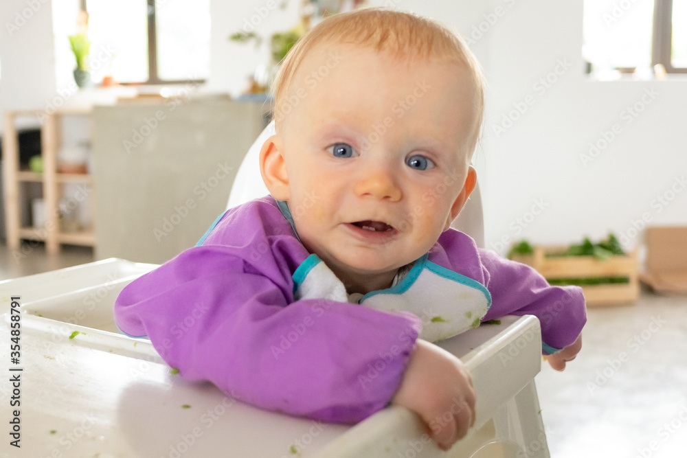 Positive blue eyed baby sitting in high chair spotted with green puree, looking at camera. Feeding or child care at home concept Positive blue eyed baby sitting in high chair spotted with green puree, looking at camera. Feeding or child care at home concept
