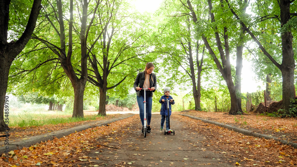 Beautiful young mother teaching her little son riding kick scooter at autumn park