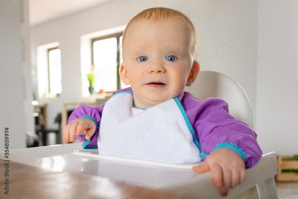 Cute red haired baby sitting in high chair for eating or feeding, looking at camera. Childhood or child care at home concept Cute red haired baby sitting in high chair for eating or feeding, looking at camera. Childhood or child care at home concept