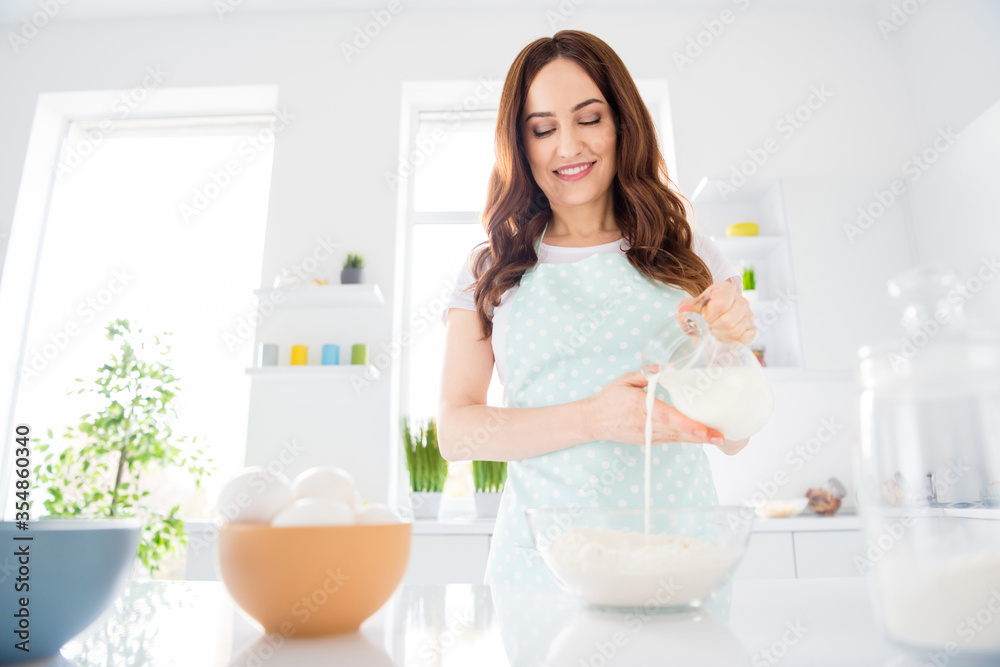 Low angle view photo of positive cheerful girl want prepare tasty cookies for her kids hold jug pour milk bowl flour wear white apron in kitchen house
