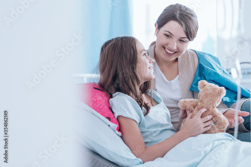 Photography Woman cheering up sick younger sister lying in hospital bed with teddy bear