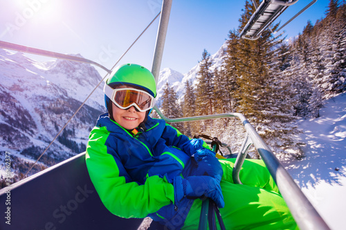Smiling little boy sit on the ski chair lift wear sport goggles and helmet look at camera over snowy forest