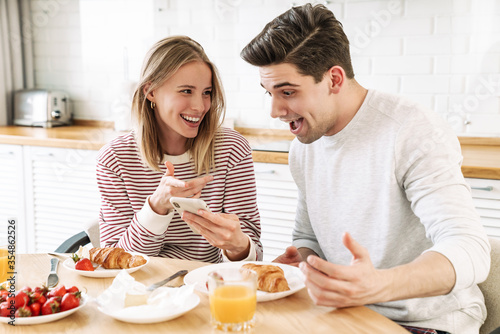 Portrait of young happy couple using smartphone while having breakfast