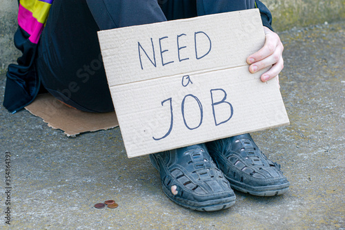 Canvas Print unemployed in old shoes holds in his hand a cardboard tablet with the inscription need a job on the ground are small coins of alms