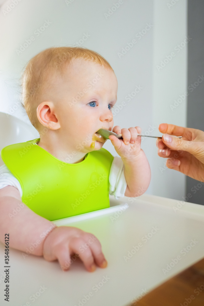 Adorable baby in green bib eating with spoon, sitting on high chair. Closeup shot. First solid food or child care at home concept Adorable baby in green bib eating with spoon, sitting on high chair. Closeup shot. First solid food or child care at home concept