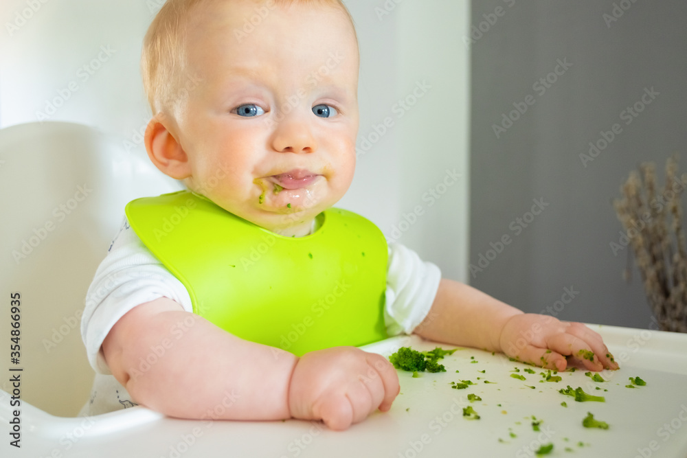 Naughty baby trying to eat broccoli vegs, posing with food spots on face, making messy on tray. Little child wearing plastic bib, sitting in highchair. First solid food or child care at home concept Naughty baby trying to eat broccoli vegs, posing with food spots on face, making messy on tray. Little child wearing plastic bib, sitting in highchair. First solid food or child care at home concept