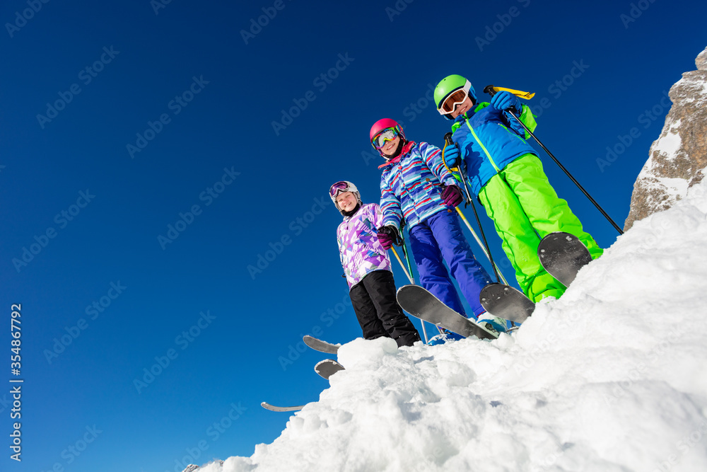 View from below of a group of three kids stand on the mountain top in snow wearing ski colorful outfit over blue sky