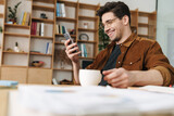 Image of joyful man smiling and using cellphone while drinking coffee