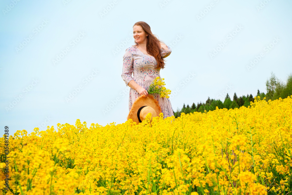 Fototapeta premium A red-haired woman in a blooming yellow field walks in flowers. Happiness, joy, youth, outdoor activities.