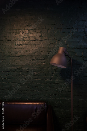 Fragment of minimalist cozy loft interior with green brick wall, floor lamp and brown leather sofa in the evening with warm lights. Place for reading and relaxing. © Daria