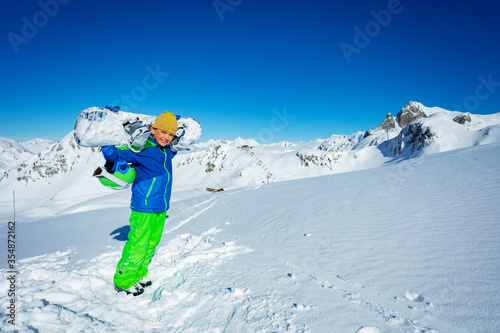 Portrait of a boy holding snowboard and casual hat on the shoulders over mountain panorama in good weather