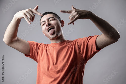 Ragazzo giovane con t-shirt arancione balla divertito facendo le corna con le mani stile rapper, isolato su sfondo grigio