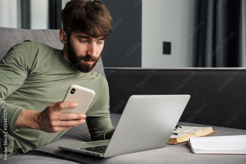 Image of young man using laptop and cellphone while lying on sofa