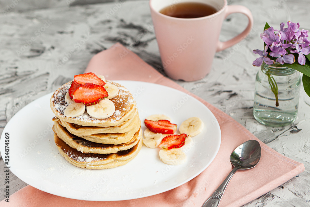 A stack of pancakes with a pink tea mug and fresh strawberries on a white plate on a light background. A small bouquet of lilacs in a glass vase stands next to the Breakfast. A stack of pancakes with a pink tea mug and fresh strawberries on a white plate on a light background. A small bouquet of lilacs in a glass vase stands next to the Breakfast.