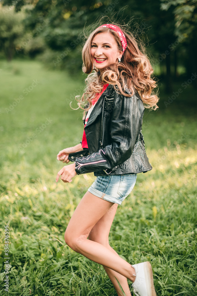 Fototapeta premium Portrait of beautiful young blonde stylish woman with long curly hair and red lipstick sitting on the green grass in rock style in short denim shorts, red headband, t-shirt, black leather jacket.