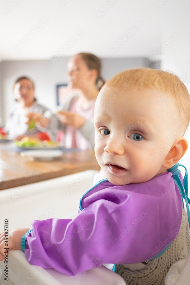 Happy red haired baby sitting in high chair with family at dining table, looking at camera. Closeup shot. Childhood or child care at home concept Happy red haired baby sitting in high chair with family at dining table, looking at camera. Closeup shot. Childhood or child care at home concept