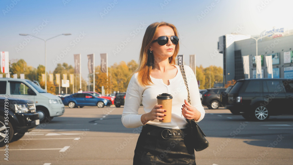 Fototapeta premium Toned portrait of young businesswoman drinking coffee from paper cup while walking on car parking to her office