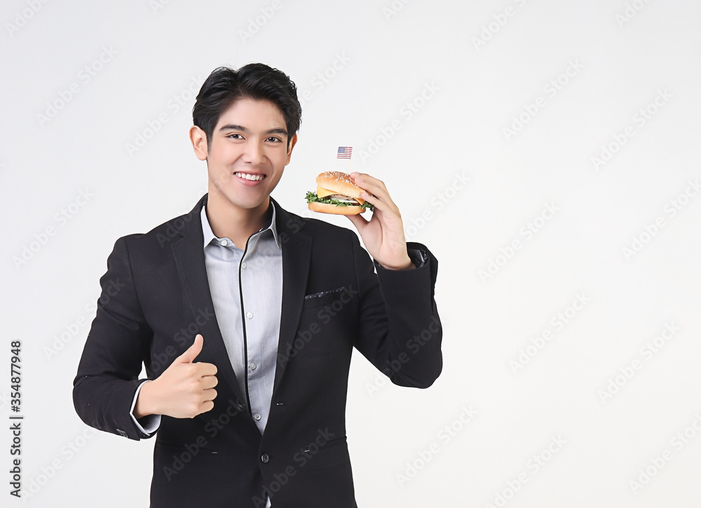 Asian young and handsome business man wearing dark suit standing on white background , holding hamburger with American flag,showing his thumb up and smiling. 4th. July , independence day concept. Asian young and handsome business man wearing dark suit standing on white background , holding hamburger with American flag,showing his thumb up and smiling. 4th. July , independence day concept.