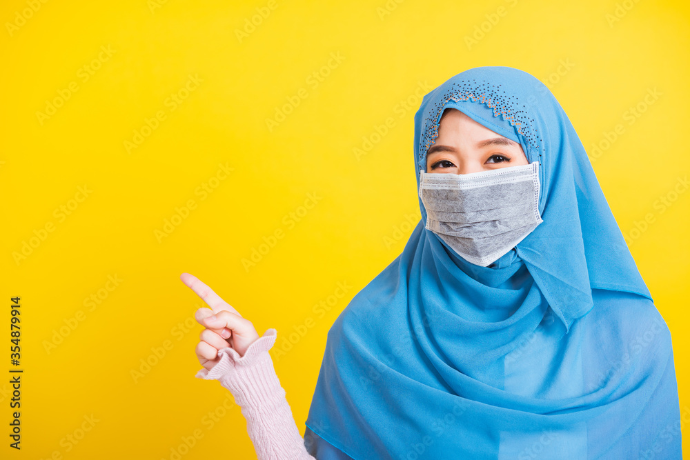 Asian Muslim Arab, Portrait of happy beautiful young woman religious wear veil hijab and face mask protective to prevent coronavirus she pointing finger side away space, isolated on yellow background