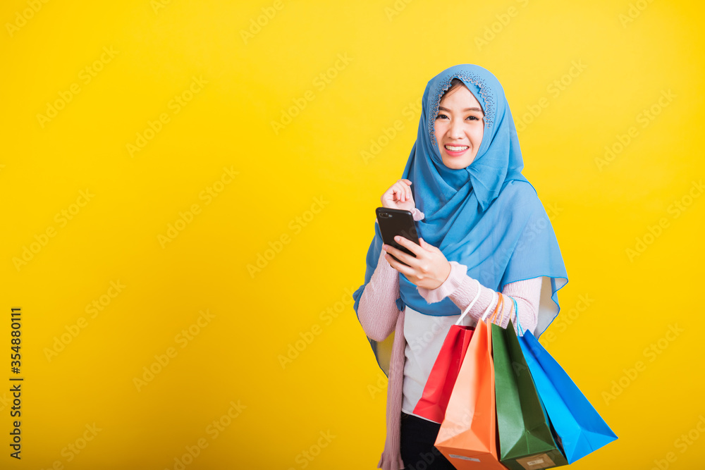 Asian Muslim Arab, Portrait of happy beautiful young woman Islam religious wear veil hijab funny smile she using smartphone on hand and hold shopping bags, studio shot isolated on yellow background