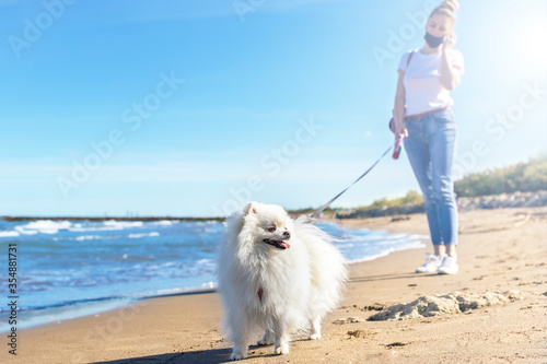 Girl with protective mask walking with dog on the beach. White pomeranian dog