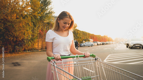 Toned portrait of beautiful smiling young girl walking with shopping trolley on car parking