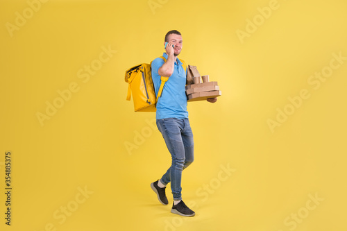 Delivery guy in blue uniform is walking talking on phone holding an order with food from restaurant and talking to customer behind his back he has delivery guy's refrigerator bag on yellow background