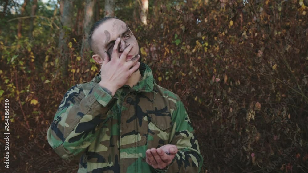 Brave soldier in camouflage smears soot strips on face at a forest before fight Brave soldier in camouflage smears soot strips on face at a forest before fight