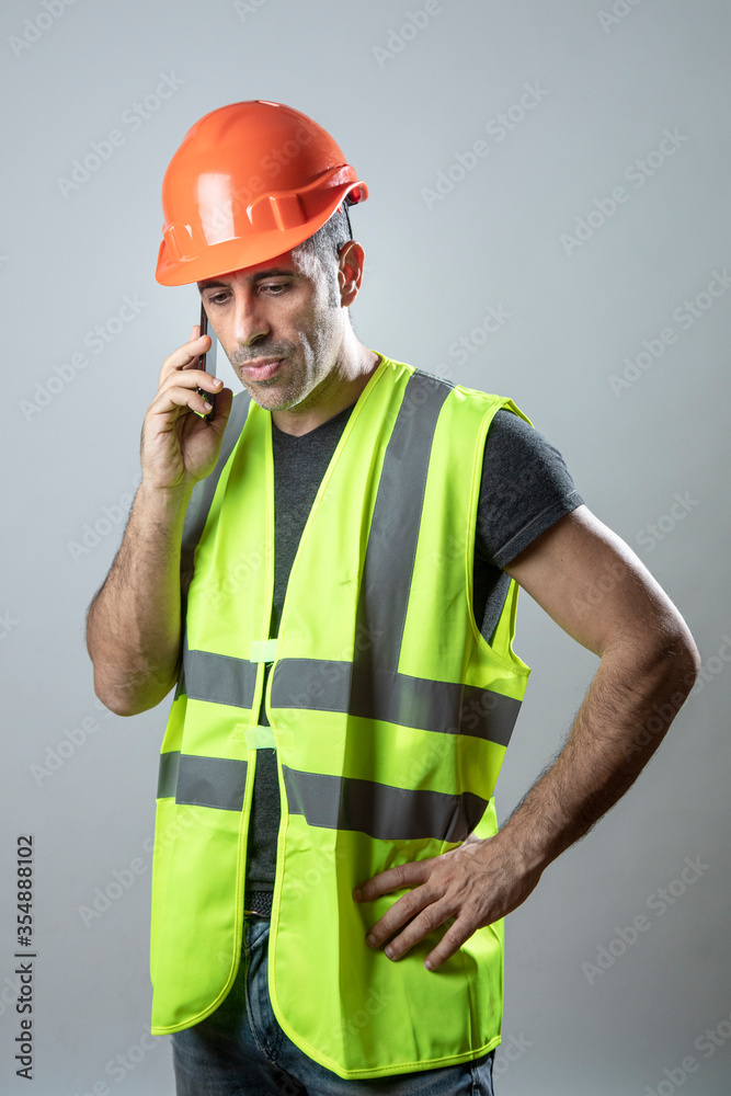 operaio con caschetto protettivo arancione e gillet giallo tenuto in mano fa una chiamata con il cellulare, isolato su sfondo grigio operaio con caschetto protettivo arancione e gillet giallo tenuto in mano fa una chiamata con il cellulare, isolato su sfondo grigio