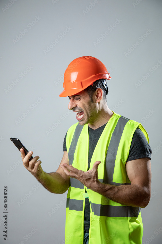 operaio con caschetto protettivo arancione e gillet giallo fa una videochiamata con il cellulare  mostrando molto disappunto, isolato su sfondo grigio