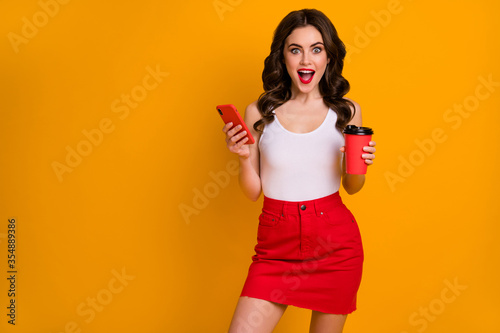 Photo of funny pretty lady hold mug coffee to-go hold telephone read new post blogger website wear casual white tank-top red skirt isolated bright yellow color background