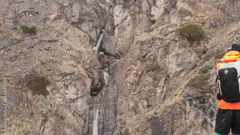 Aerial view of a young male photographer with a camera in his hands stands on a high rock in a gorge and photographs an alpine waterfall during a snowstorm and snowfall in spring. Aerial view of a young male photographer with a camera in his hands stands on a high rock in a gorge and photographs an alpine waterfall during a snowstorm and snowfall in spring.