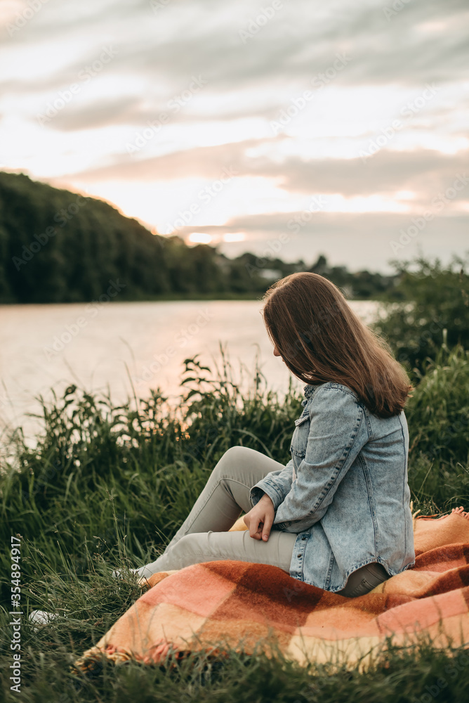 Young white beautiful sad woman sitting on plaid in denim, jeans clothes on nature in summer near lake at sunset. 