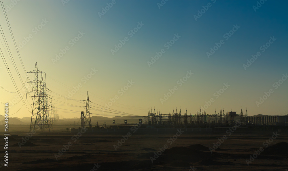 A power station far out in the desert in southern Egypt. The electricity pylons soar into the sky. In the background are the mountains in front of the desert.